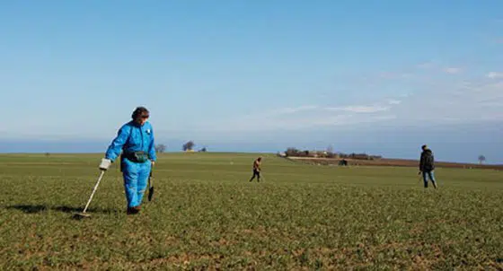 Tre personer, der går på mark med metaldetektorer. Marken er grøn, og der er blå himmel