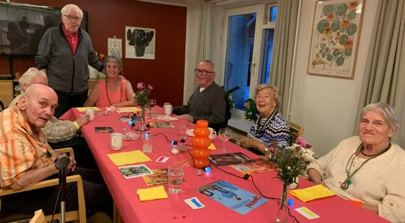 Seks ælndre mænd og kvinder sidder rundt om et langbord med rød dug. De smiler til kameraet. En mand står op, for enden af bordet. Han holder venstre hånd på skulderen på den kvinder, der sidder for bordenden.