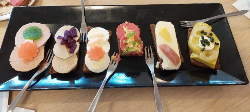 Seks madder med pålæg ligger på en sort tallerken med fem gafler