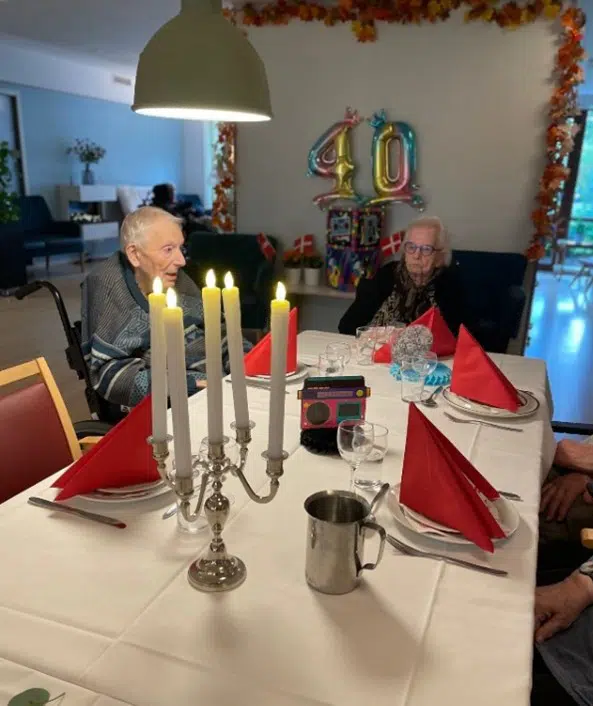 To ældre mennesker sidder ved et bord med hvid dug og røde servietter og en femarmet lysestage i forgrunden. I baggrunden hænger to tal balloner, som symboliserer, at der fejres 40-års fødselsdag