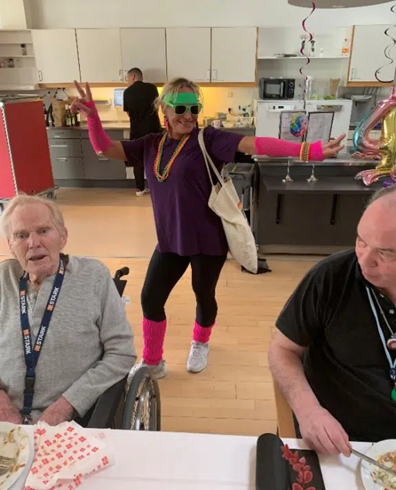 To ældre mænd sidder ved et fødselsdagsdækket bord. Bag dem står en kvinde med sorte gamacher, pink ben- og håndledsvarmere, halskæde i selvlysende farver, skriggrønt pandbånd og solbriller.