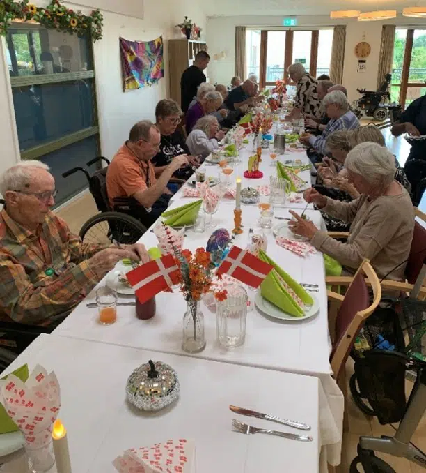 Mange ældre mænd og kvinder sidder ved at fødselsdagsdækket langbord. Der er flag på bordet og på servietterne.