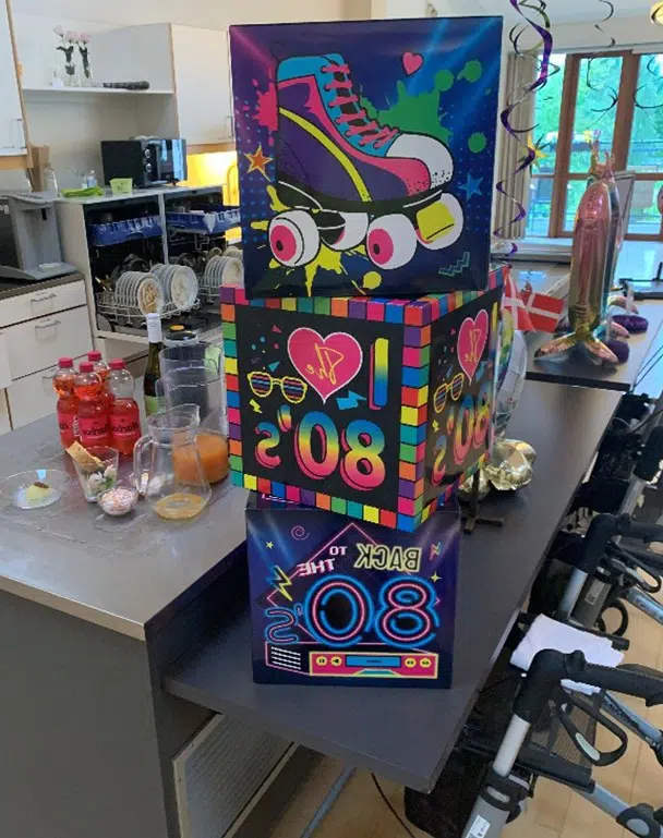 På et køkkenbord står tre kasser i et stabel. På den øverste er der et stort billedet af en rulleskøjte, på den midterste tern fra en rubicskube og solbriller. På den nederste en pladespiller.