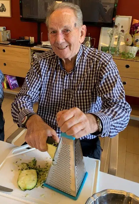 En ældre mand river en stor squash på et rivejern. Han smiler til fotografen