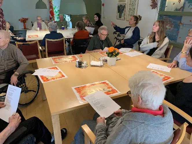 Ved borde sidder ældre og unge mennesker. De synger 