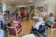 En flok beboere og personale sidder på stole med hulahopringe i hænderne