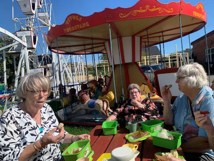 Tre beboere fra Egegården sidder ved en bænk og spiser madpakker. I baggrunden ses en karrusel