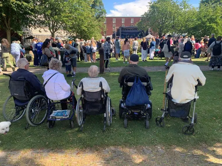 Fem beboere fra Egegården sidder og ser frem på en scene til Gladsaxedagen. En stor menneskemængde af tilskuere ses også