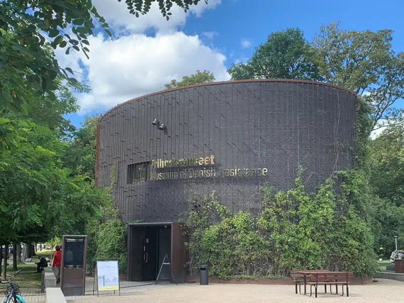 Frihedsmuseet set ude fra. Der vokser klematis og andre slyngplanter opad murene. Museet er en moderne oval bygning i mørkegrå mursten. Der står Frihedsmuseet på både dansk og engelsk i "guld" bogstaver på faseden