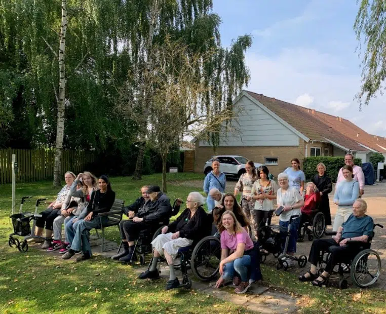 En stor flok beboere fra Egegården, elever fra en ungdomsskole og en medarbejder ses siddende på bænke. Nogle enkelte står op bagved