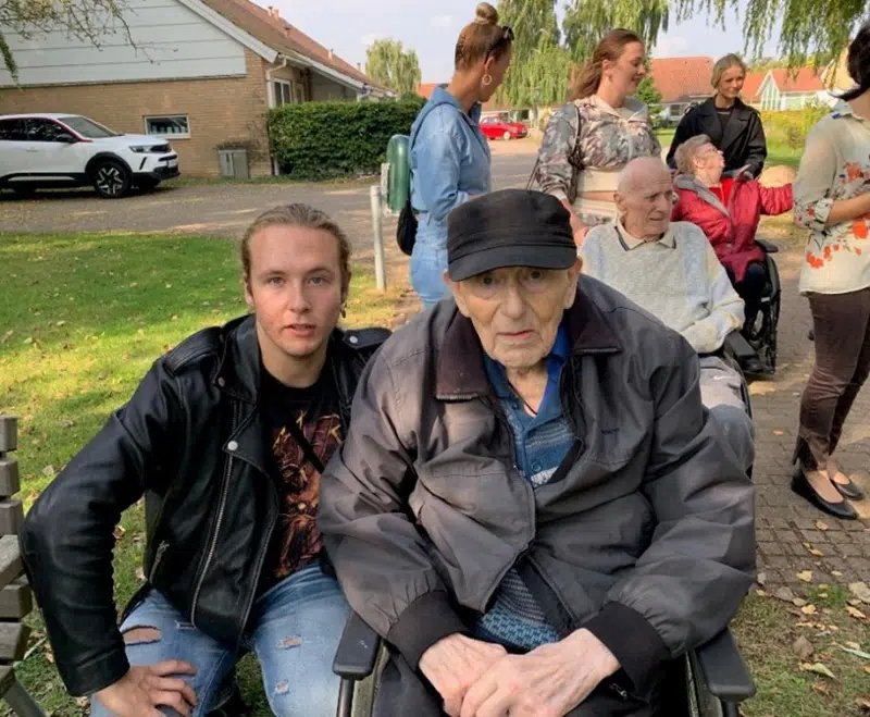 En beboer fra Egegården sidder i kørestol og ved siden af sidder en elev fra ungdomsskolen på hug. I baggrunden ses andre beboere og elever