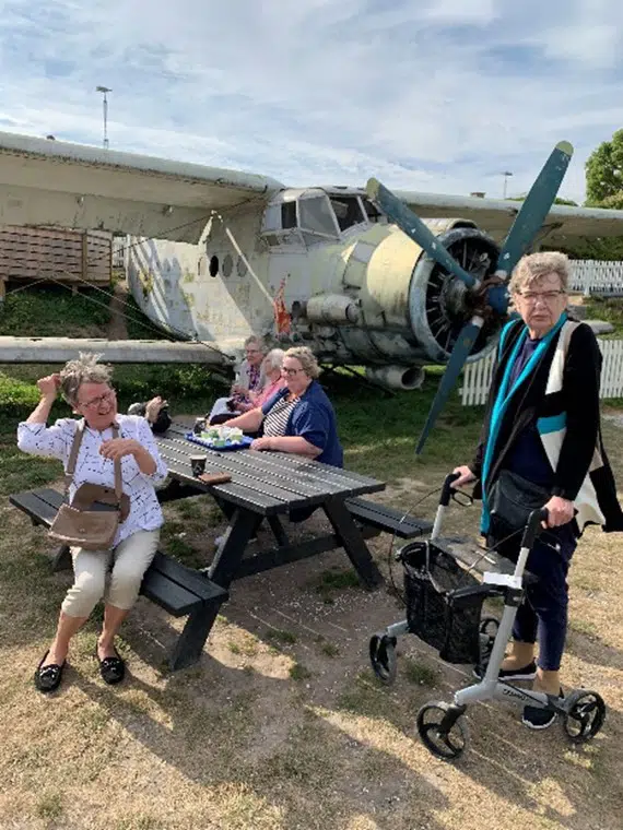 Nogle beboere fra Egegården sidder på en bænk. En beboer står op med en rollator. I baggrunden ses et gammelt propelfly