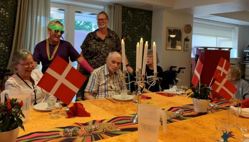 Nogle ældre mennersker sidder ved et bord med gul dug i nervøst velour. Bagved står to kvinder, hvoraf den ene er iklædt tøj inspireret af 80’erne. 