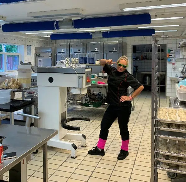 En køkkenmedarbejder står i køkkenet. Hun smiler til kameraet, har skrigpink benvarmere på og skriggrønne solbriller.