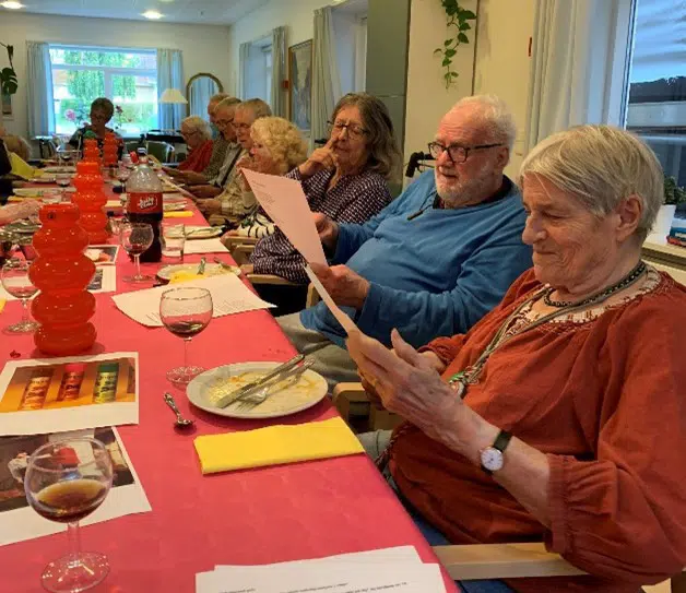 10 ældre mennesker sidder omkring et lanbord dække med rød dug. De har service foransig og sidder og kigger på et papir. Damen tættest på fotografen har et smil på læben, mens hun kigger på papiret.