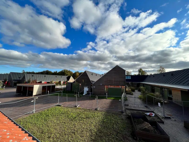 Et moderne boligområde med lave bygninger med mørke tage og træfacader. Foran bygningerne er der grønne områder, nyanlagte stier og afspærringer, der indikerer igangværende anlægsarbejde. Himlen er blå med spredte skyer, og området fremstår lyst og indbydende.