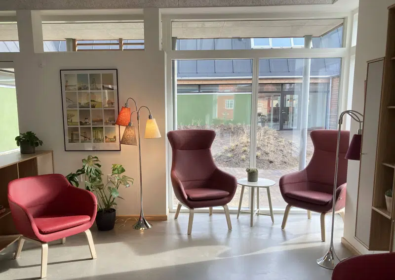 Et lyst og indbydende opholdsrum med store vinduer, der giver udsigt til en gårdhave. I rummet står tre moderne stole med rødt betræk, et lille rundt bord med en plante, flere grønne stueplanter og to gulvlamper med farvede lampeskærme. Rummet fremstår hyggeligt og velegnet til afslapning eller samtale.