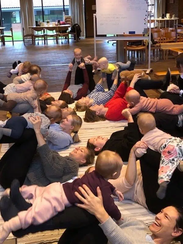 10 kvinder ligger i rundkreds med benene mod hinanden. Deres knæ er bøjede og på deres underben ligger babyer. 