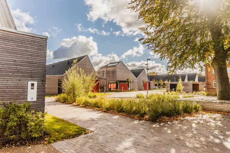 Et moderne boligområde med træbeklædte huse, grønne buske og træer samt en brolagt sti. Solen skinner gennem trækronerne, og himlen er delvist dækket af skyer. Området fremstår lyst, indbydende og velplejet.