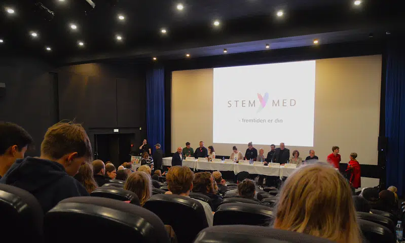 En fyldt biografsal med politikere på scenen, ovenfor står der "stem med" på det store lærred.