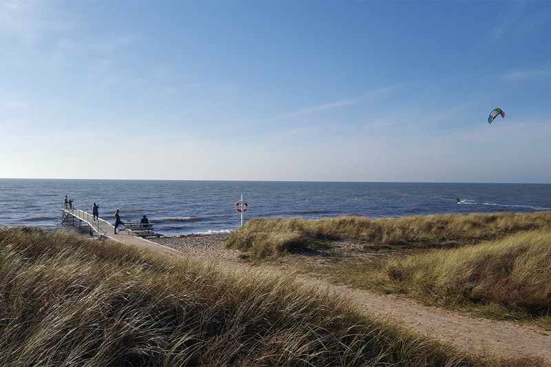 Foto af stranden med mennesker på badebro og kitesurfer i vandet